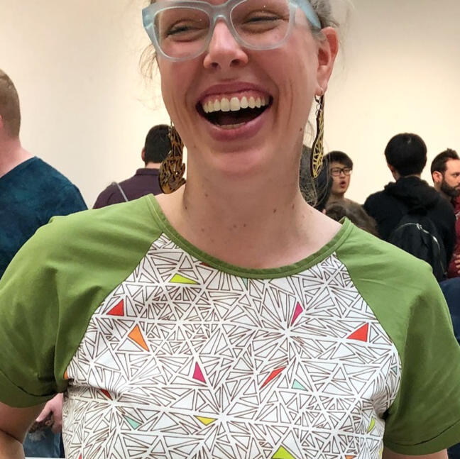 woman laughing, wearing blue glasses and a shirt with green sleeves and a printed front fabric with triangle drawings